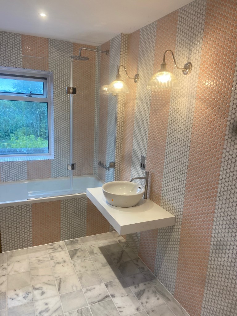 Modern bathroom installation with feature penny tiles, wall-mounted basin, bath shower combination and designer lighting completed in West Sussex.